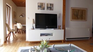 En moderne stue med et fladskærms-tv på et hvidt tv-møbel, trægulv og et spisebord i baggrunden.