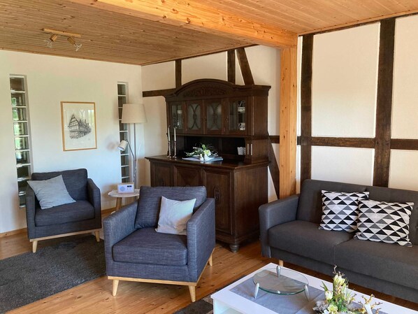 Uma sala de estar acolhedora com teto de madeira, um armário de madeira escura, dois sofás cinzas e uma pequena mesa branca com uma luminária.