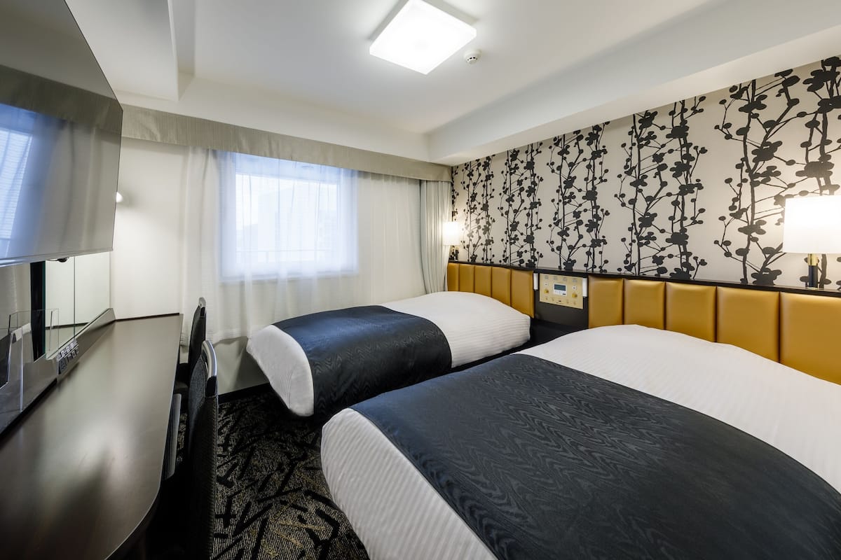 Hotellihuone, jossa on kaksi sänkyä, työpöytä, taulutelevisio, ikkuna, jossa on läpikuultavat verhot, ja kukkakuviotapetti.