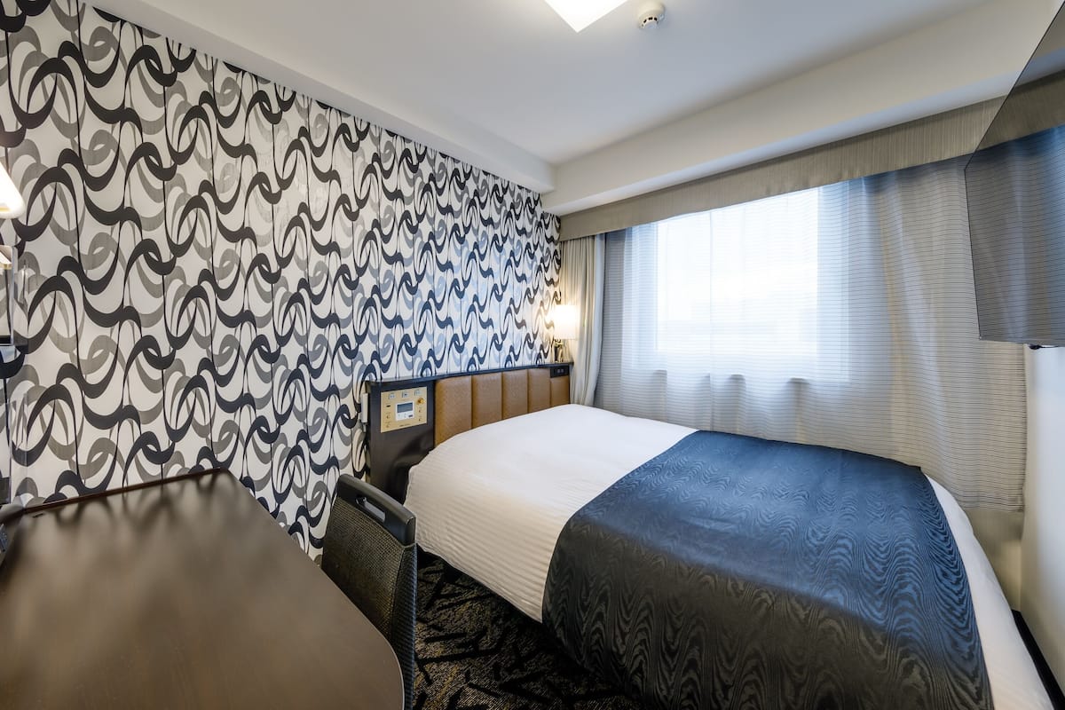 Hotellihuone, jossa on suuri sänky, työpöytä ja tuoli. Huoneen seinillä on kuvioitu tapetti ja ikkunassa on verhot.