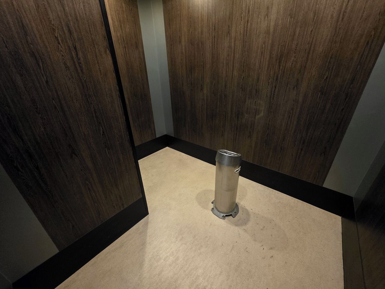 Ein Zimmer mit Holzboden, einem Metallmülleimer und einem beigen Teppich.