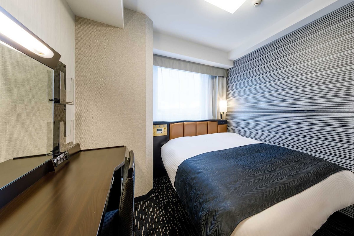 Hotellihuone, jossa on sänky, yöpöytä, peili, ikkuna verhoilla ja raidallinen tapetti yhdellä seinällä.
