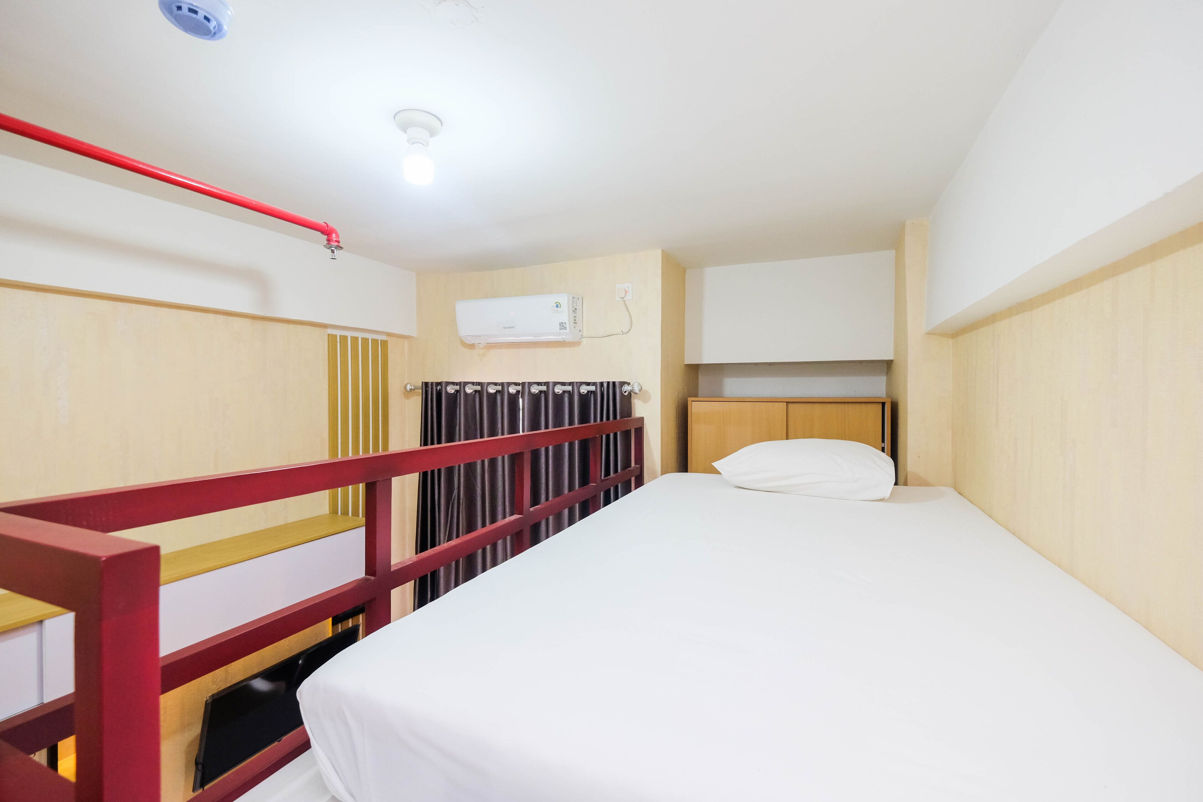 Habitación individual con una cama de madera, cabecera de madera, baranda roja, colchón blanco y almohada blanca. Incluye aire acondicionado montado en la pared y una lámpara en el techo.