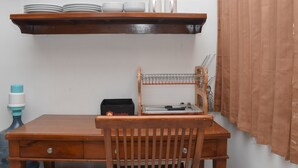 Una mesa de madera con una silla, un estante con platos y tazas, un bolso de viaje y un escurridor de metal con utensilios.