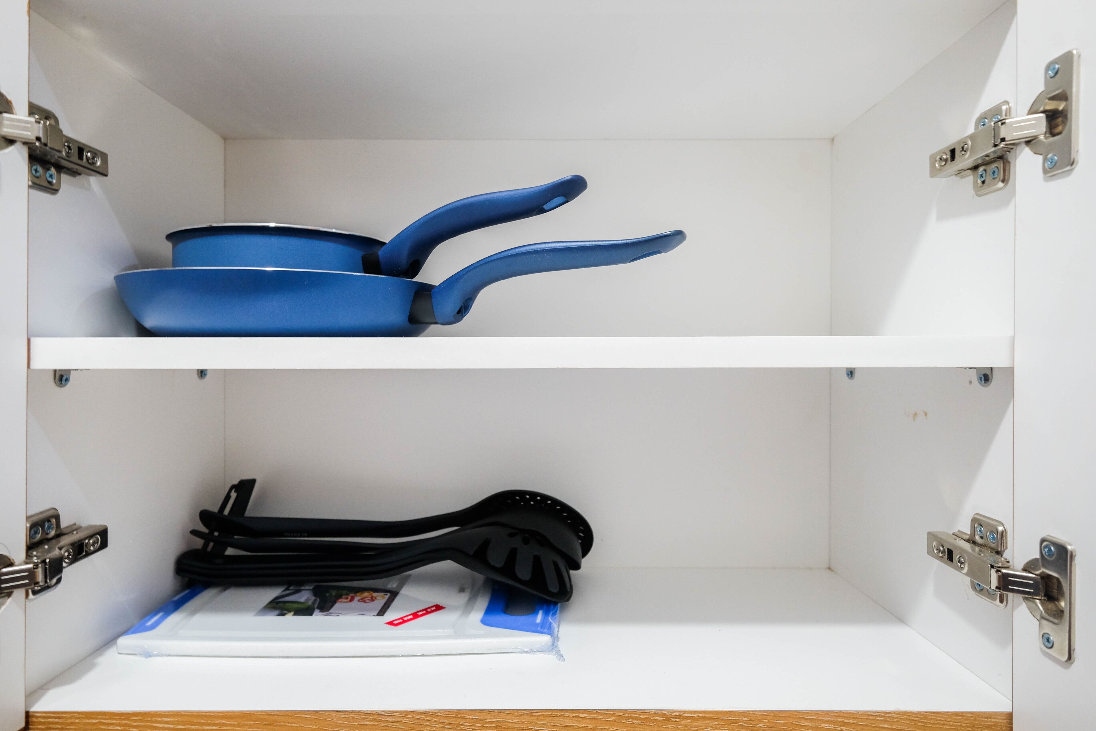 A white cabinet with two shelves; the top shelf holds a blue pan and a black spatula, and the bottom shelf contains a white tray with a black spatula.