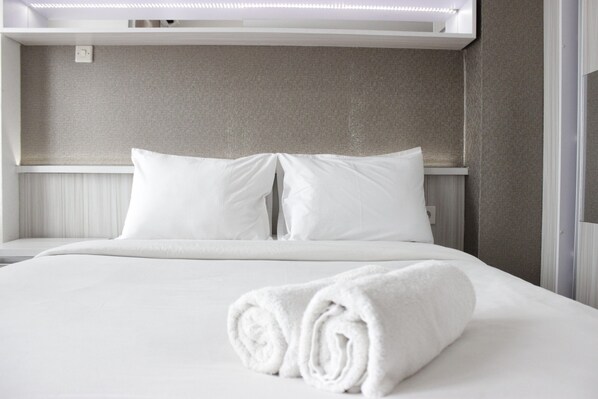 A neatly made bed with white linens and a rolled towel, a light switch, and a textured wall. - Pleasurable Studio Room Near Unpad At Taman Melati Jatinangor Apartment (Hegarmanah)