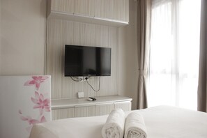 A hotel room with a bed, a flat-screen TV mounted on the wall, a bedside table, and a window with curtains. - Pleasurable Studio Room Near Unpad At Taman Melati Jatinangor Apartment (Hegarmanah)