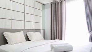 A neatly made bed with white linens, a headboard with a grid pattern, and a window with curtains. - Comfort 1Br At Casa De Parco Apartment (Pagedangan)