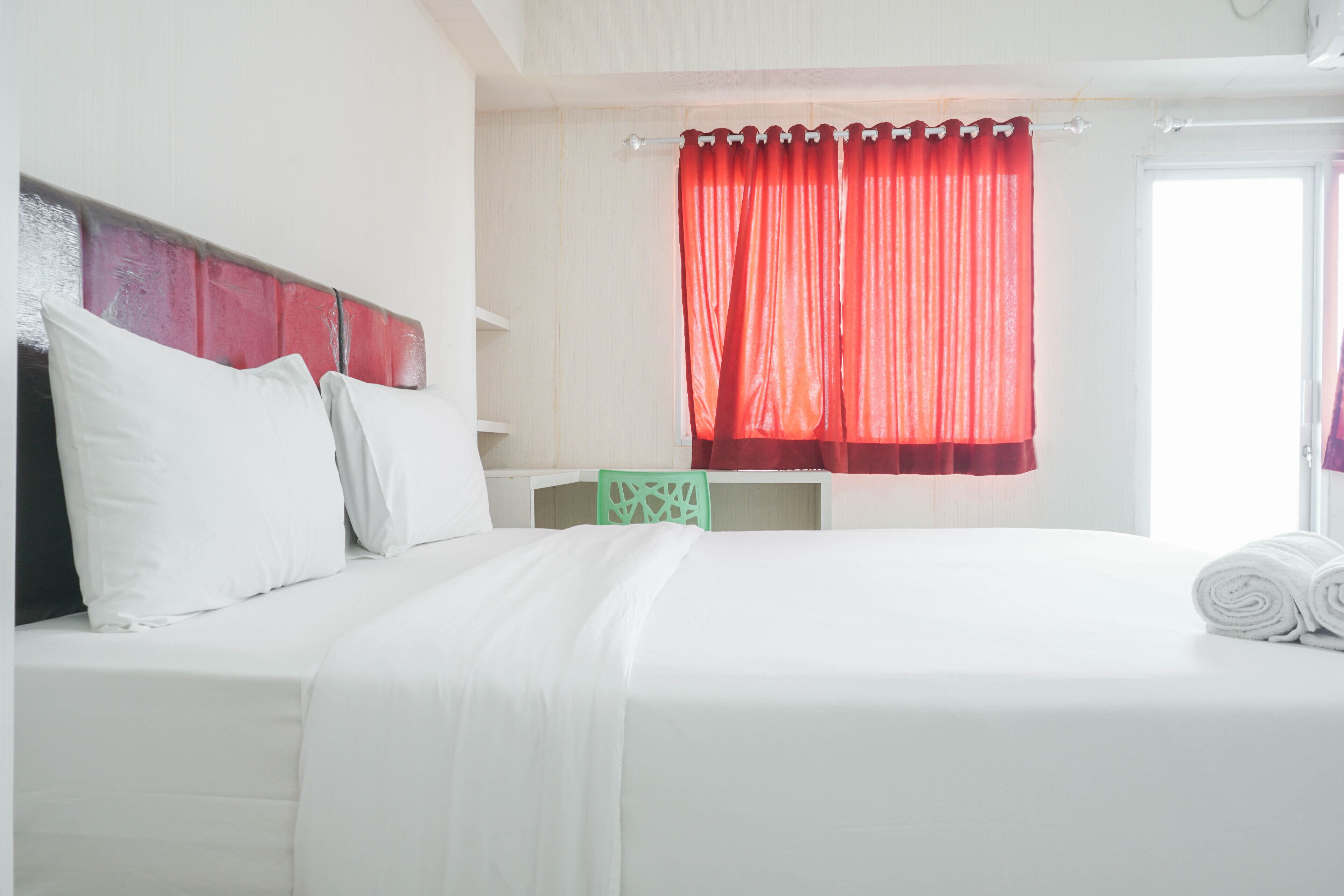 Uma cama bem arrumada com lençóis brancos, uma cadeira verde e uma cortina vermelha em um quarto de hotel.