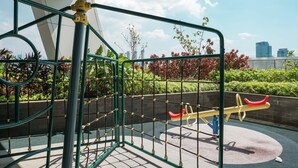A playground with a seesaw and climbing structures, surrounded by greenery and a cityscape in the background.