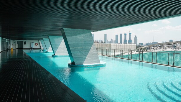 A modern indoor swimming pool with a city skyline view, featuring a sleek architectural design with a wooden deck and a metal railing.