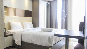 A modern hotel room with a large bed, a bedside table, a small desk, and a view of the outside through a window. - Nice And Fancy Studio Room At Menteng Park Apartment (Jakarta)