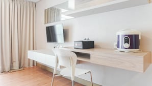 A modern hotel room with a wooden floor, a white chair, a flat-screen TV mounted on the wall, a small table with a kettle, and a window with curtains. - Comfortable And Nice Studio Room Apartement At H Residence (Jakarta)