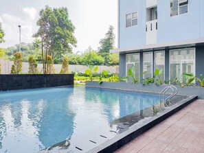 A modern swimming pool with a wooden deck, surrounded by lush greenery and a contemporary building with large windows.