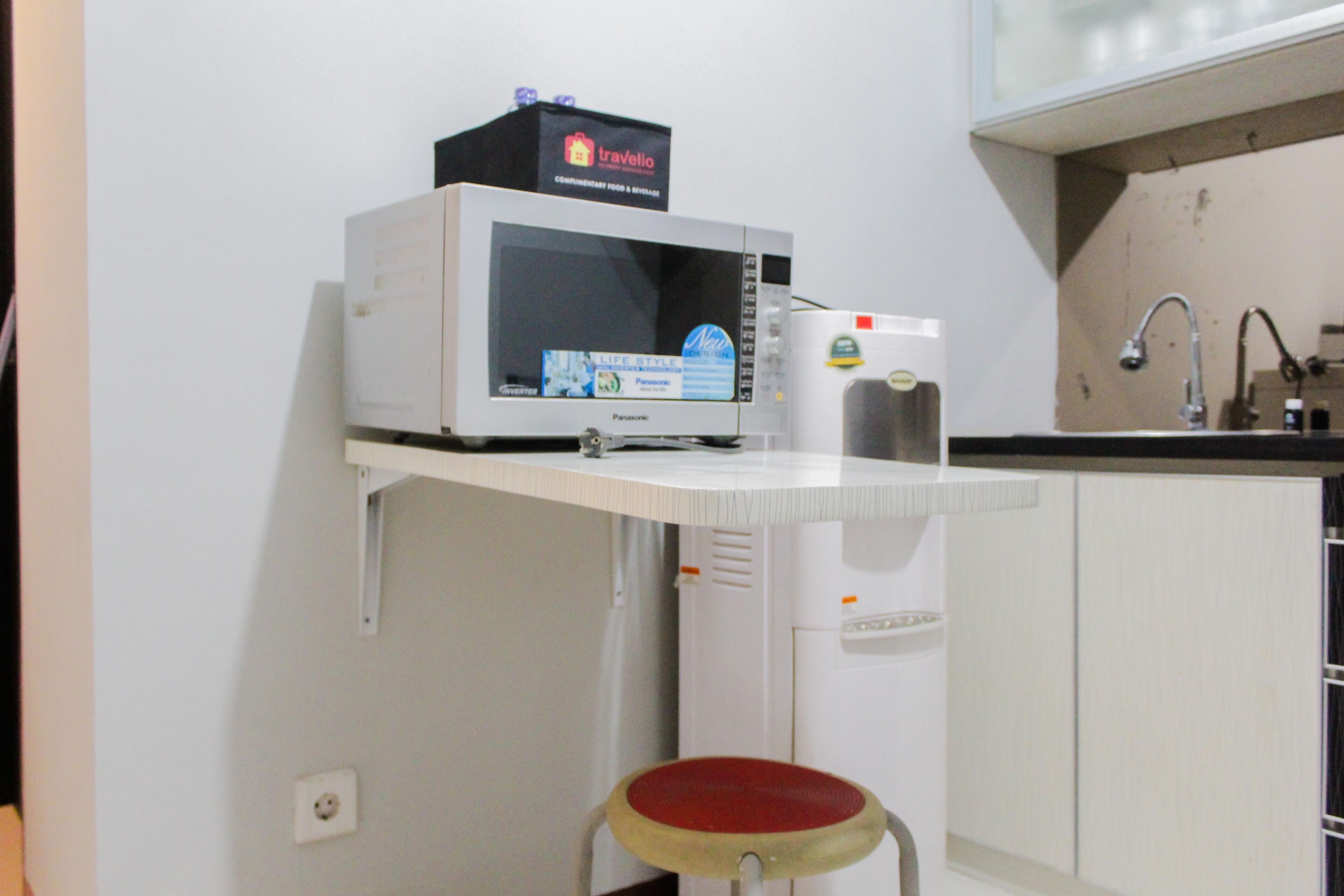 A kitchen with a microwave, a refrigerator, a sink, and a stool.