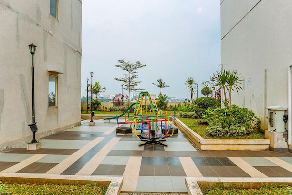 A playground with a climbing structure and a slide, surrounded by a paved area with a lamp post and a small garden with palm trees.