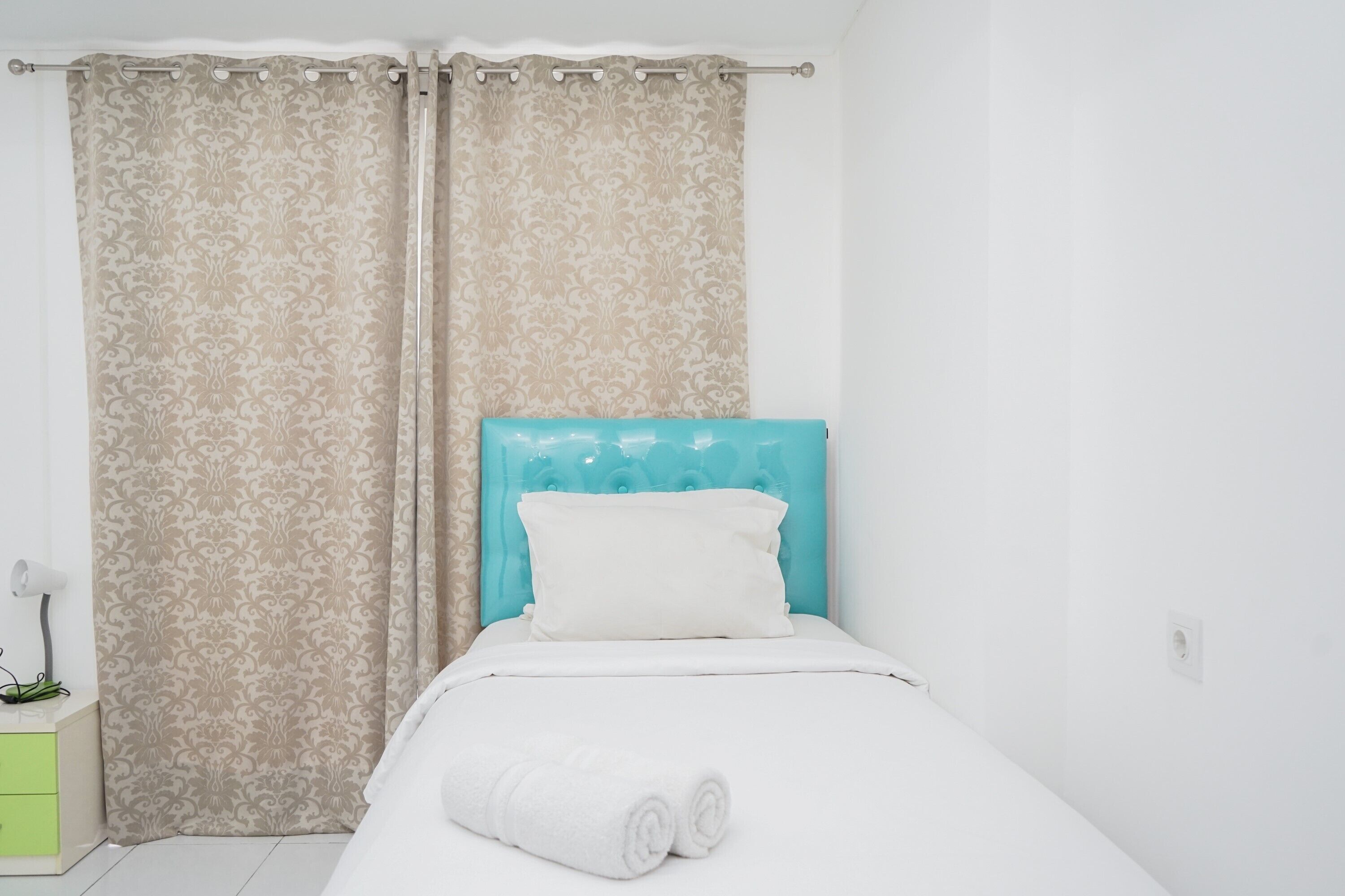 Un lit bien fait, avec une couette blanche et des serviettes blanches pliées, une tête de lit turquoise et une table de chevet verte sur laquelle se trouve une lampe.