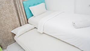 A neatly made bed with white linens and a blue headboard, a rolled white towel on the bed, and a patterned curtain in the background. - Simply 1Br At Casa De Parco Apartment (Pagedangan)