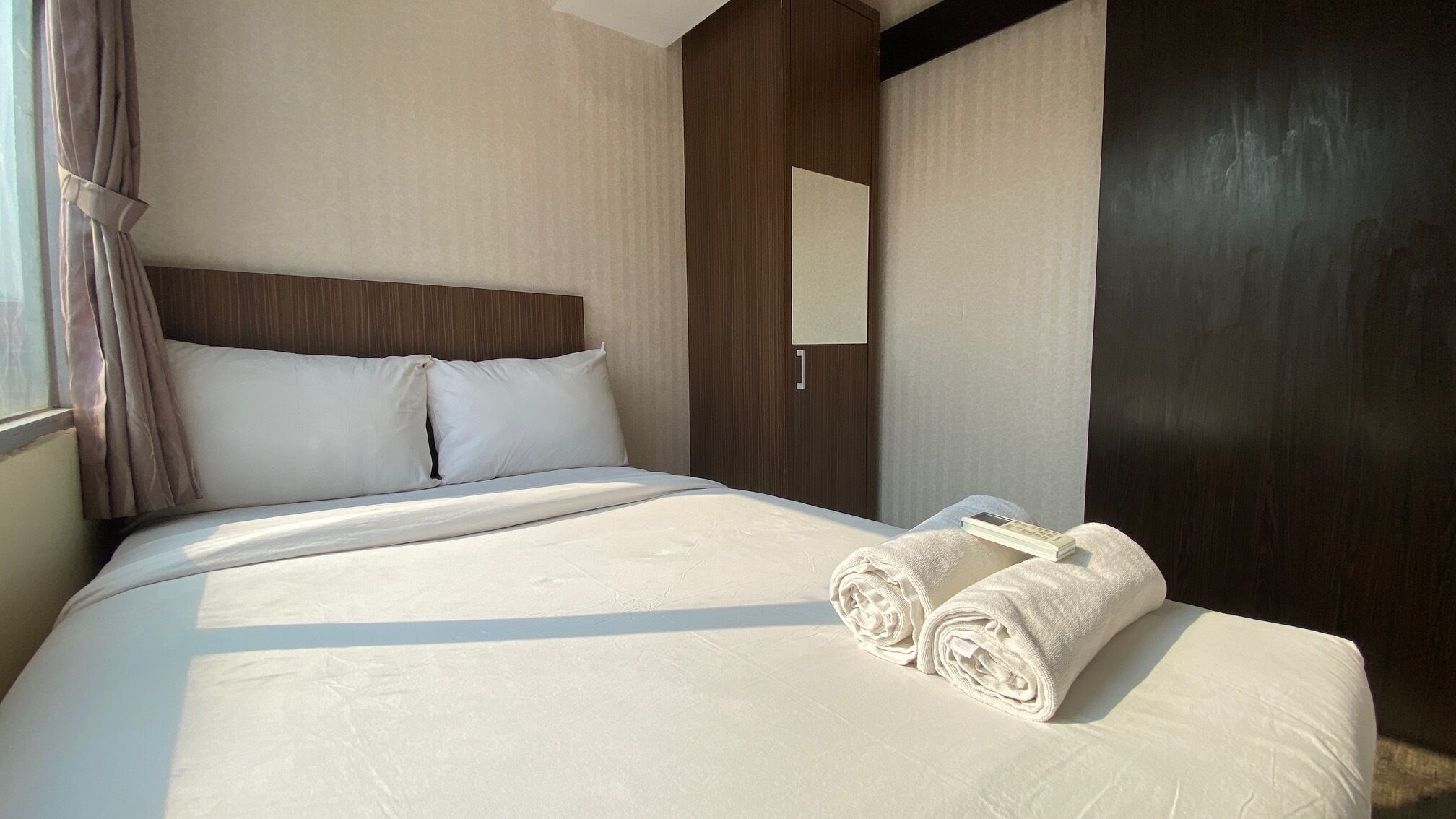 Une chambre d’hôtel avec un grand lit, une tête de lit en bois, une table de chevet avec une télécommande et une serviette soigneusement pliée.