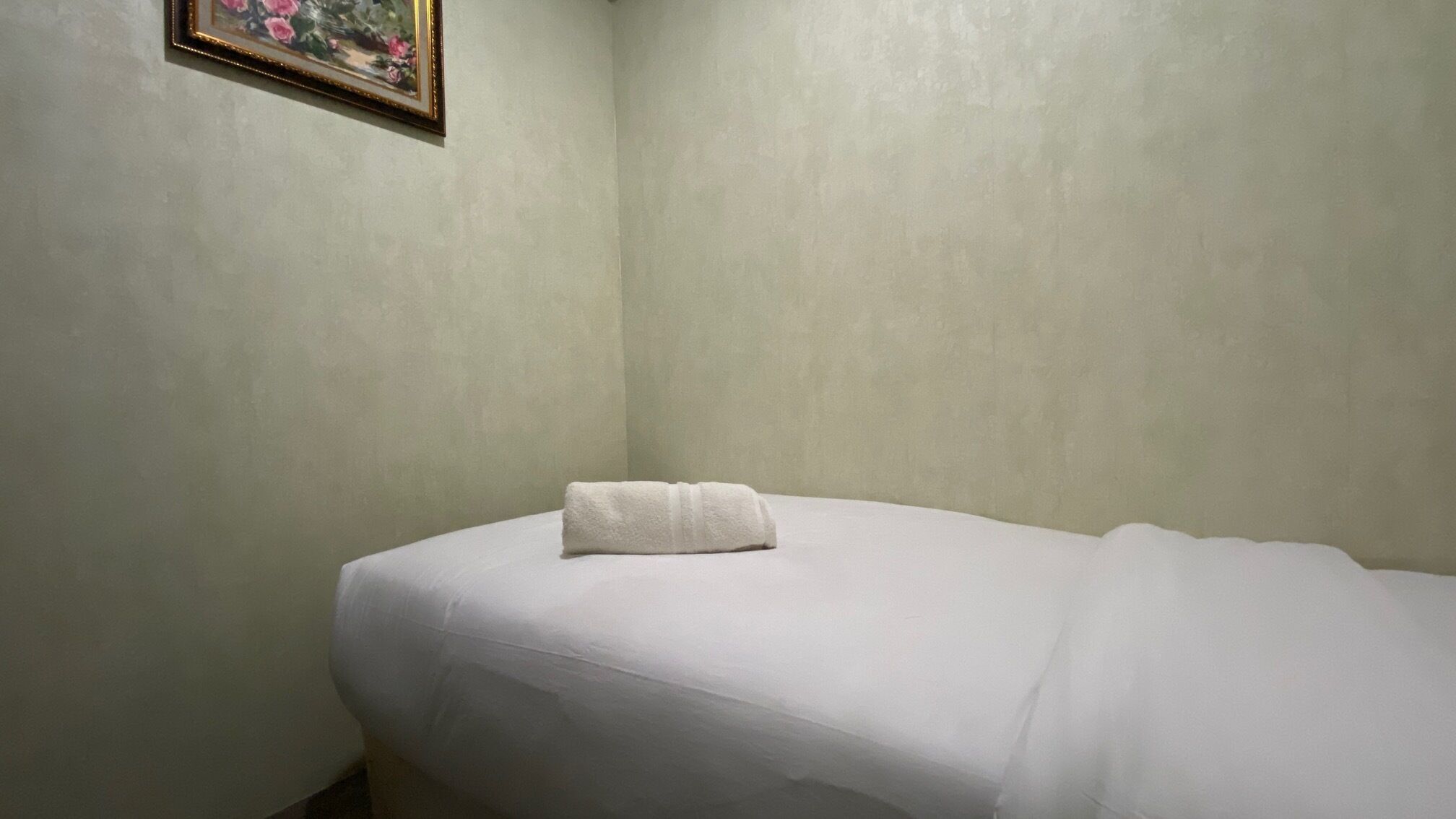 Ein kleines Zimmer mit einem Einzelbett, einer schlichten Wand und einem gerahmten Bild.