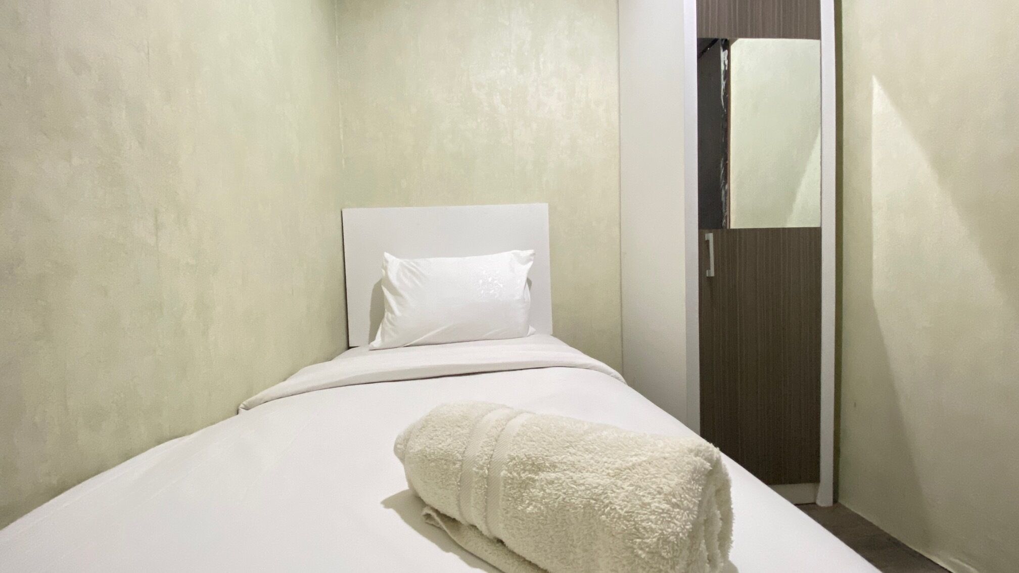 A small, neatly made bed with a white headboard and a single white pillow. A rolled white towel is placed at the foot of the bed. The room has plain walls and a door with a towel hanging on the doorknob.