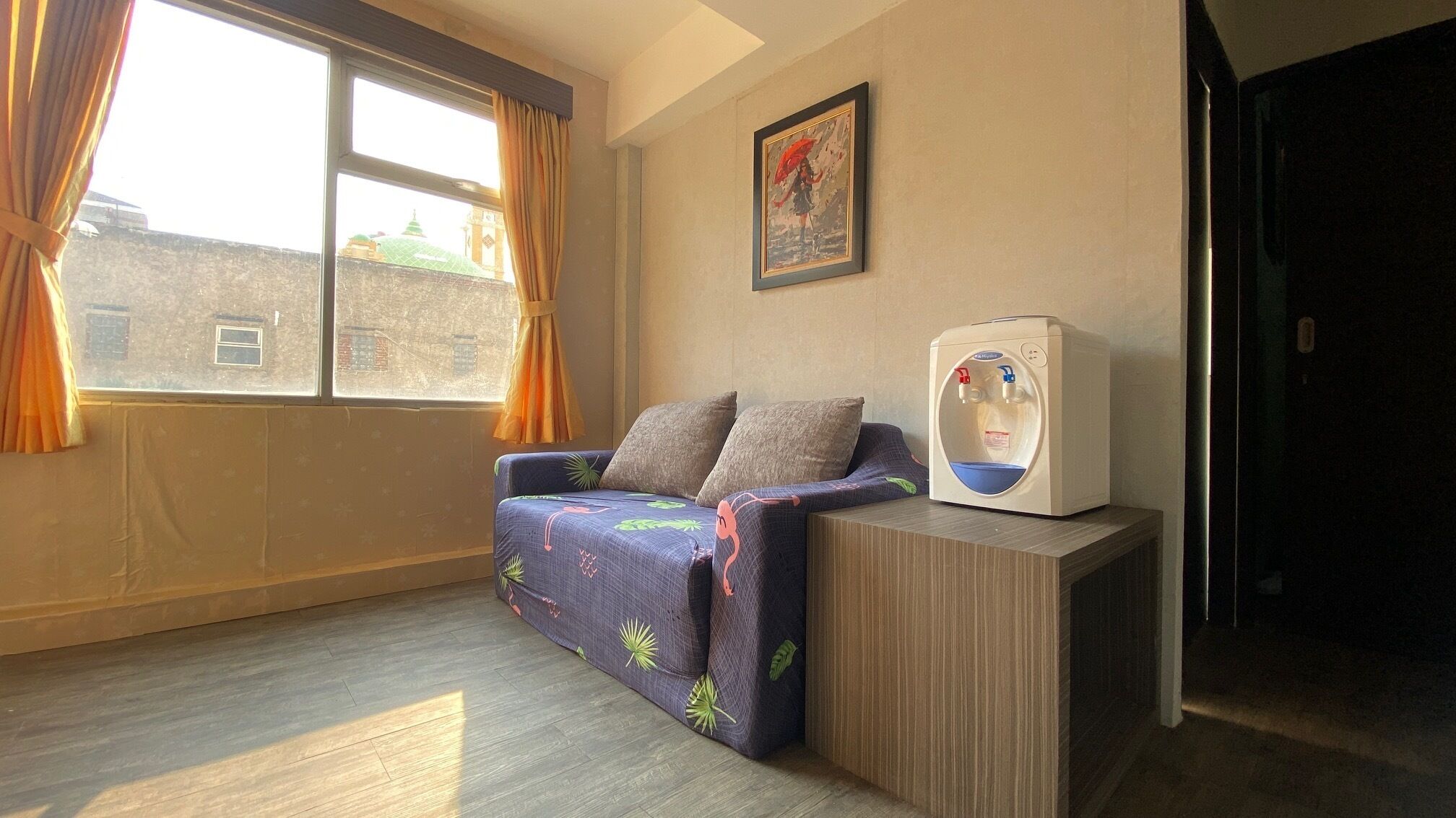 A room with a sofa, a water dispenser, a window with curtains, and a framed picture on the wall.
