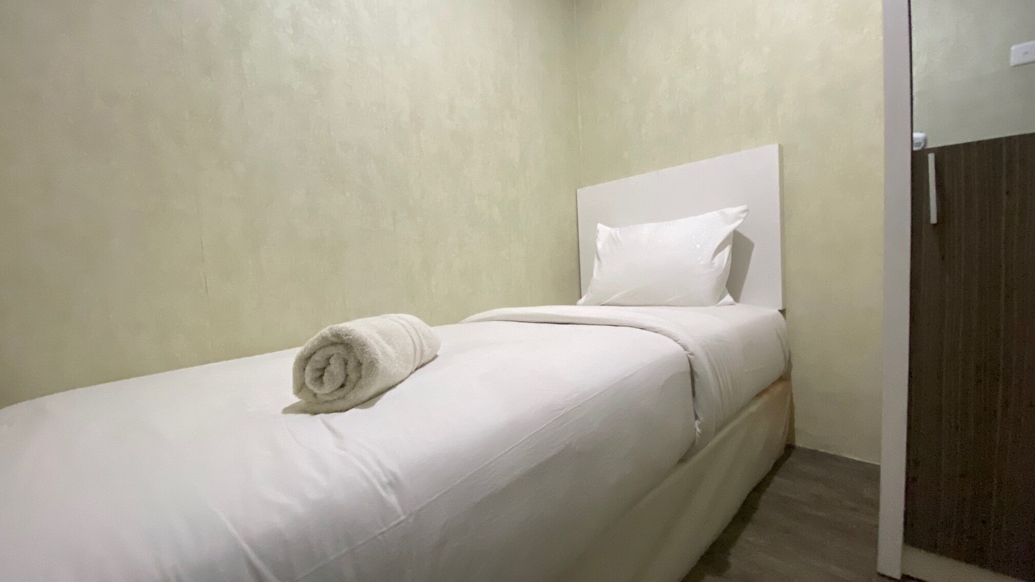 Een enkel bed met wit beddengoed en kussens, een opgerolde handdoek op het bed, en een houten deur aan de rechterkant.