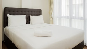 A neatly made bed with white linens and a rolled white towel, a dark upholstered headboard, and sheer curtains on a window. - Homey 2Br At Asatti Apartment (Pagedangan)