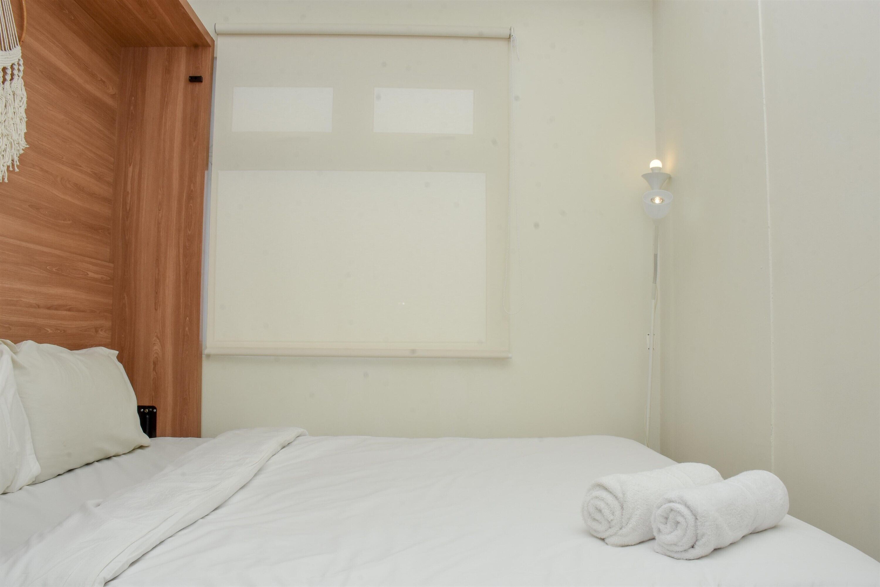 A hotel room with a wooden headboard, a bed with white bedding, a wall-mounted light, and a window with blinds.