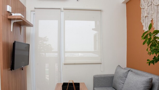 A modern living room with a grey sofa, a wooden coffee table with a plant, a flat-screen TV mounted on the wall, and a sliding glass door leading to a balcony. - 2Br Yotayori Home At Green Pramuka City Apartment (Jakarta)
