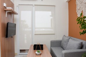 A modern living room with a grey sofa, a wooden coffee table with a plant, a flat-screen TV mounted on the wall, and a sliding glass door leading to a balcony.
