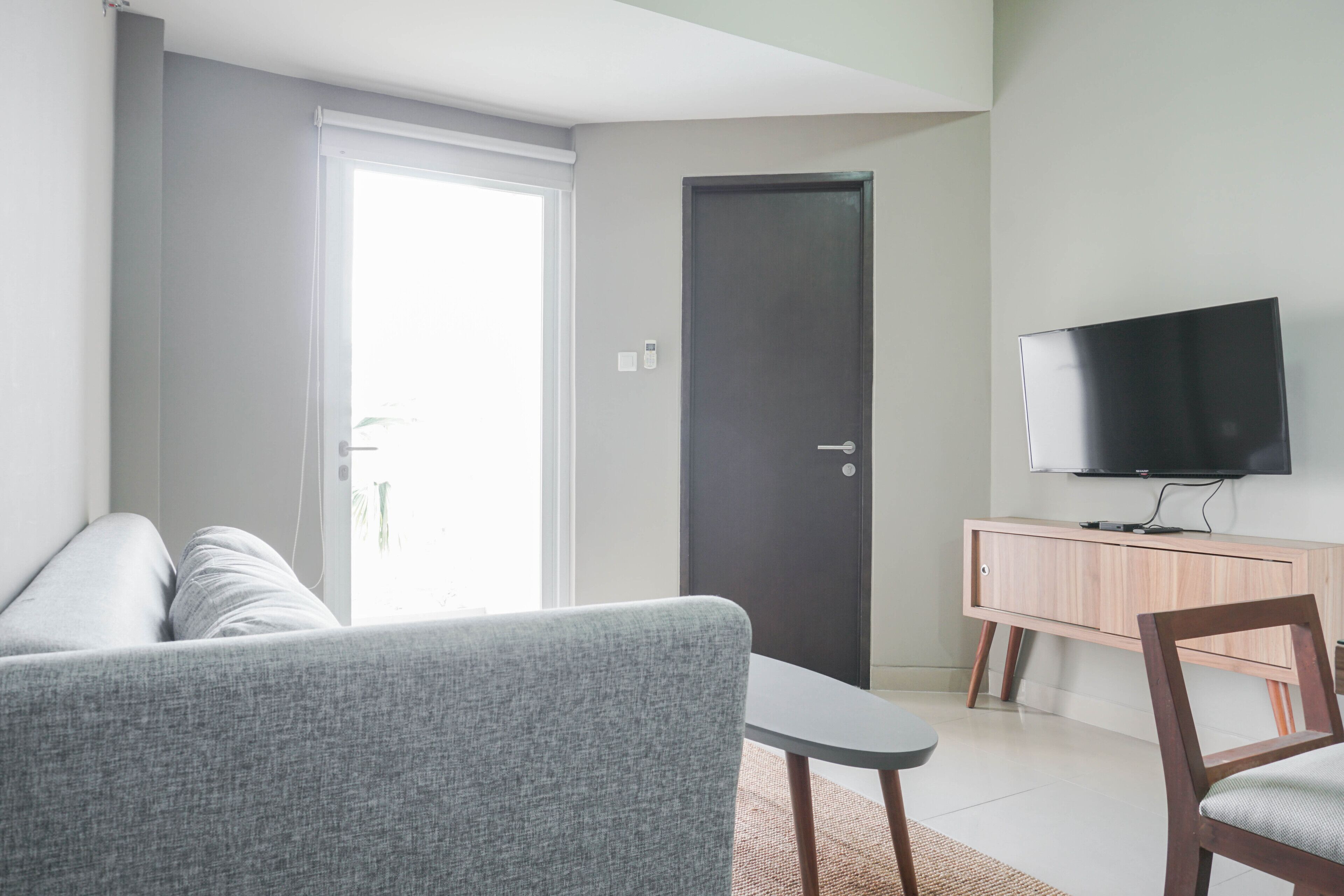 A modern living room with a grey sofa, a small table, a wooden chair, a TV on a wooden cabinet, and a door.