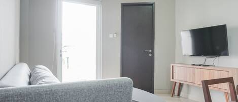 A modern living room with a grey sofa, a small table, a wooden chair, a TV on a wooden cabinet, and a door.