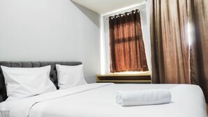Ein ordentlich bezogenes Bett mit weißen Leinen und einem aufgerollten weißen Handtuch, ein dunkel gepolstertes Kopfteil und ein Fenster mit braunen Vorhängen.