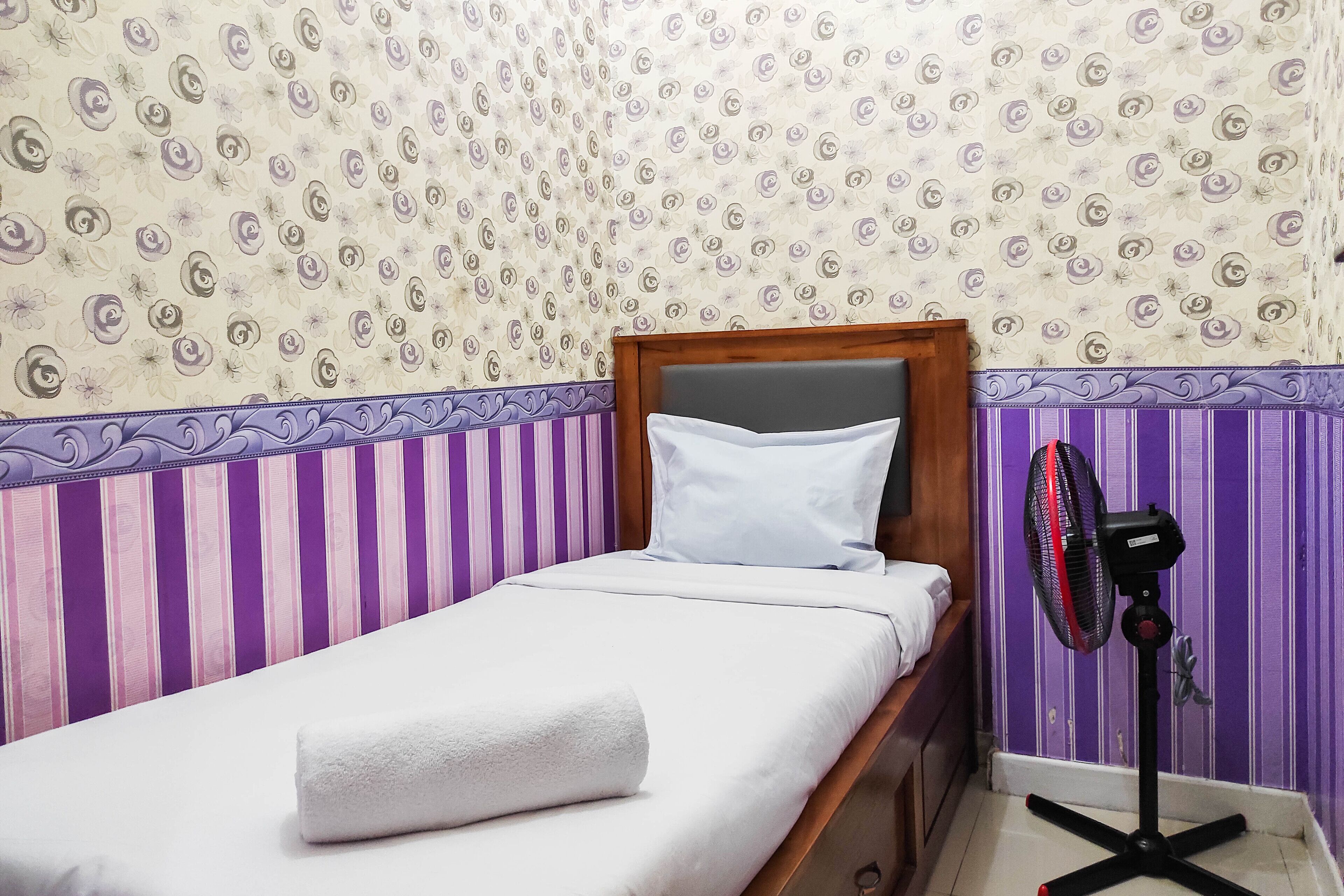 Un lit simple avec un oreiller blanc et une serviette blanche enroulée, une tête de lit en bois et un ventilateur noir posé contre un mur recouvert d’un papier peint à rayures violettes et blanches.