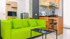 A modern living room with a bright green sofa, a small black table, and a kitchen area with wooden cabinets, a tiled backsplash, and a built-in oven. - Alluring 2Br At Bassura City Apartment Near Mall (Jakarta)
