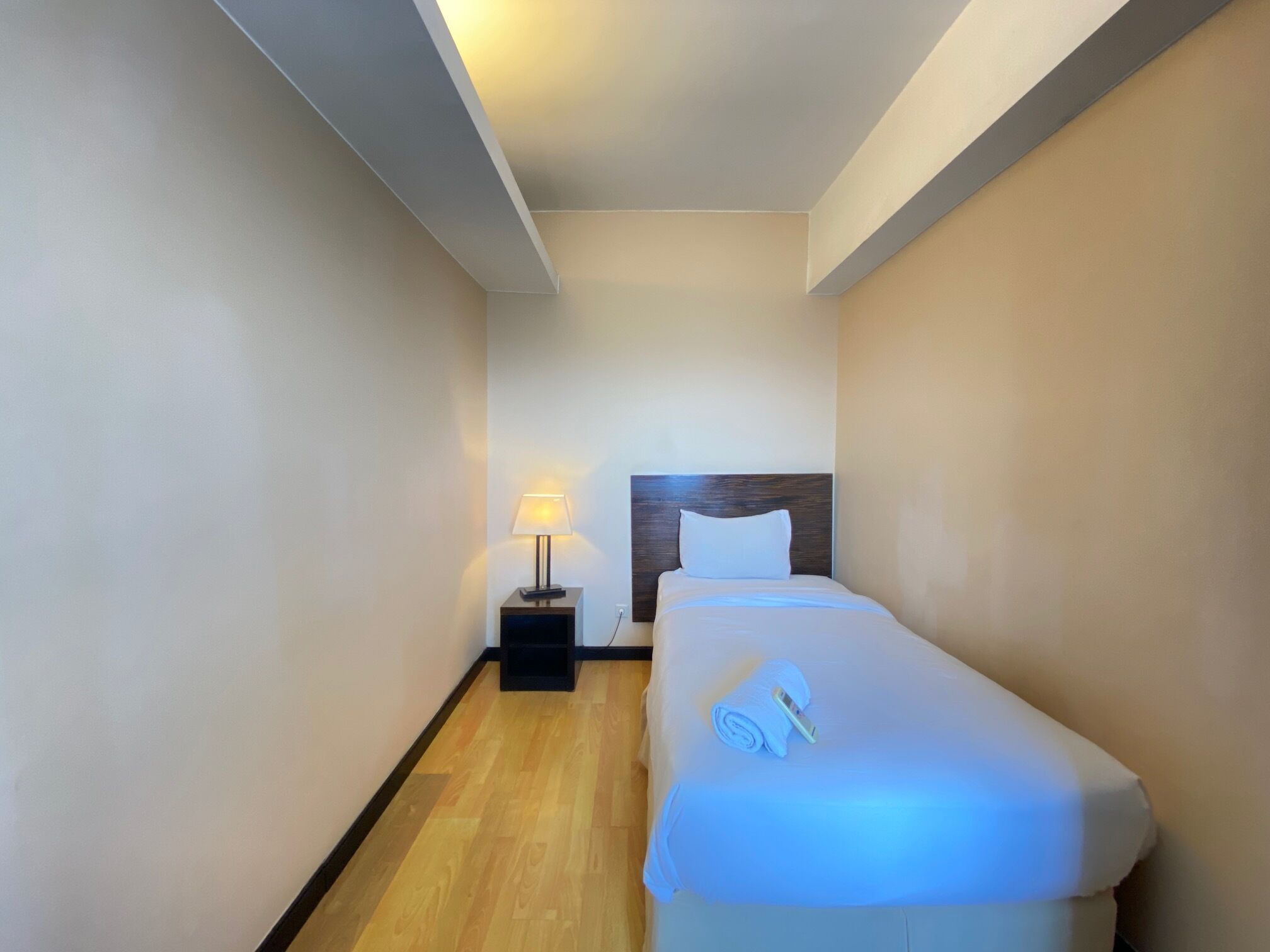 A small, narrow hotel room with a single bed, a nightstand with a lamp, and a wooden floor.