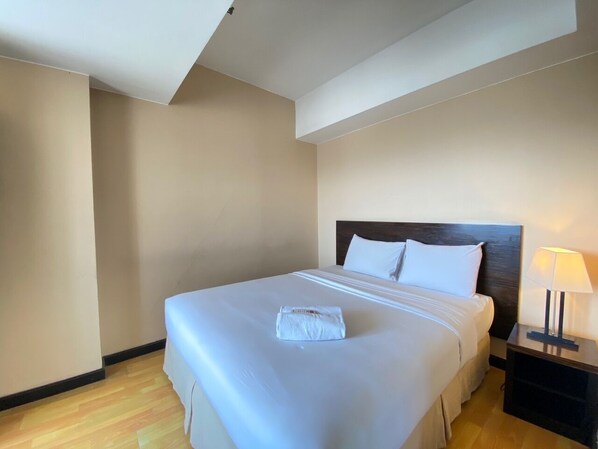 A neatly made bed with a folded towel, a wooden headboard, and a bedside table with a lamp. - Gorgeous & Classic 2Br At Braga City Walk Apartment (Bandung)