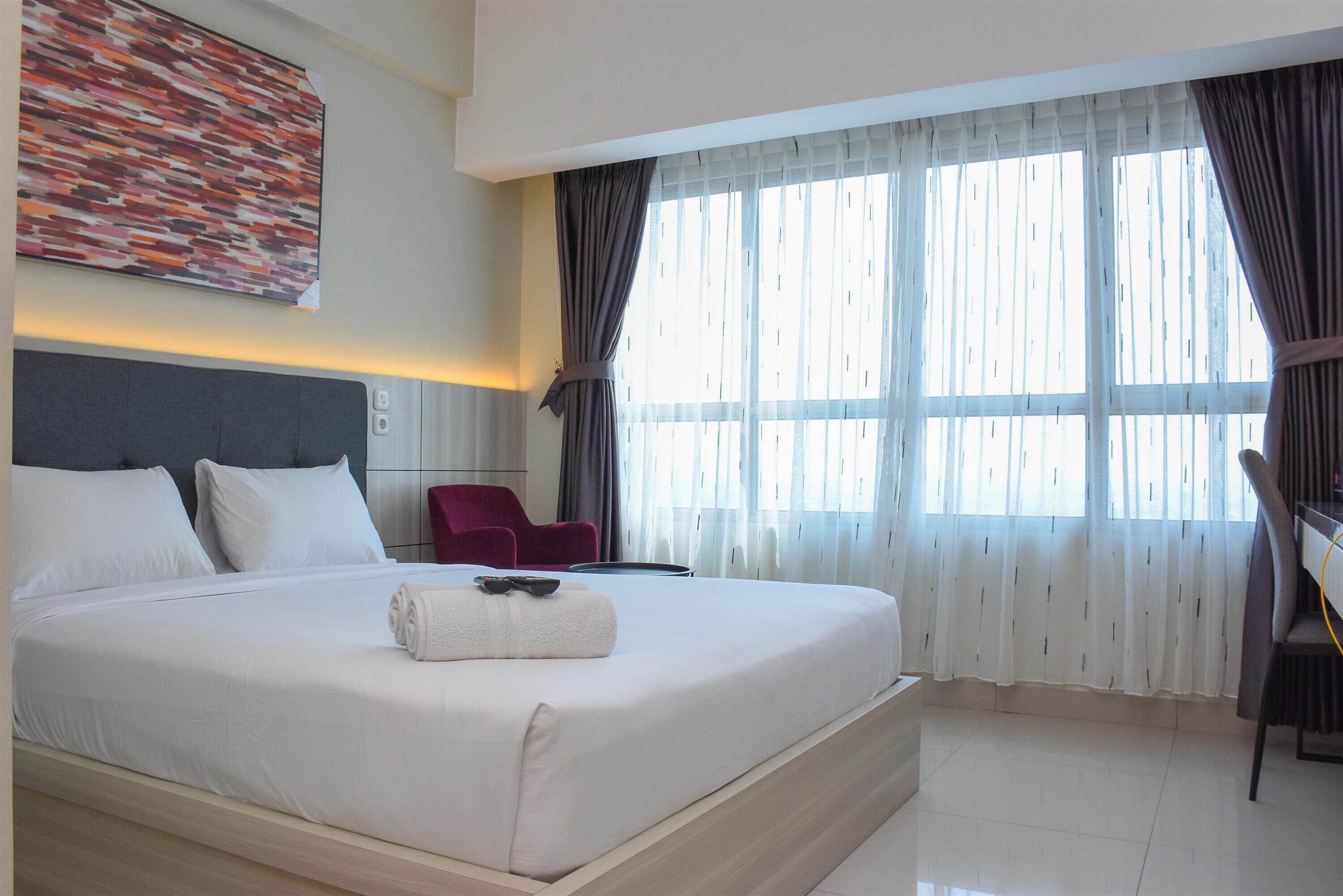 A hotel room with a large bed, a red chair, a desk, and a window with curtains.