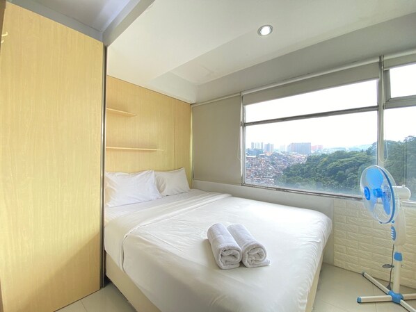 Relax in a spacious room with a large bed, a standing fan, and a window offering a city view.