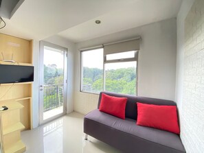 A modern living room with a grey sofa, red cushions, a flat-screen TV on a wooden shelf, and a balcony with a city view. - Comfortable 2Br Apartment Ac In Living Room At The Jarrdin Cihampelas (Bandung)