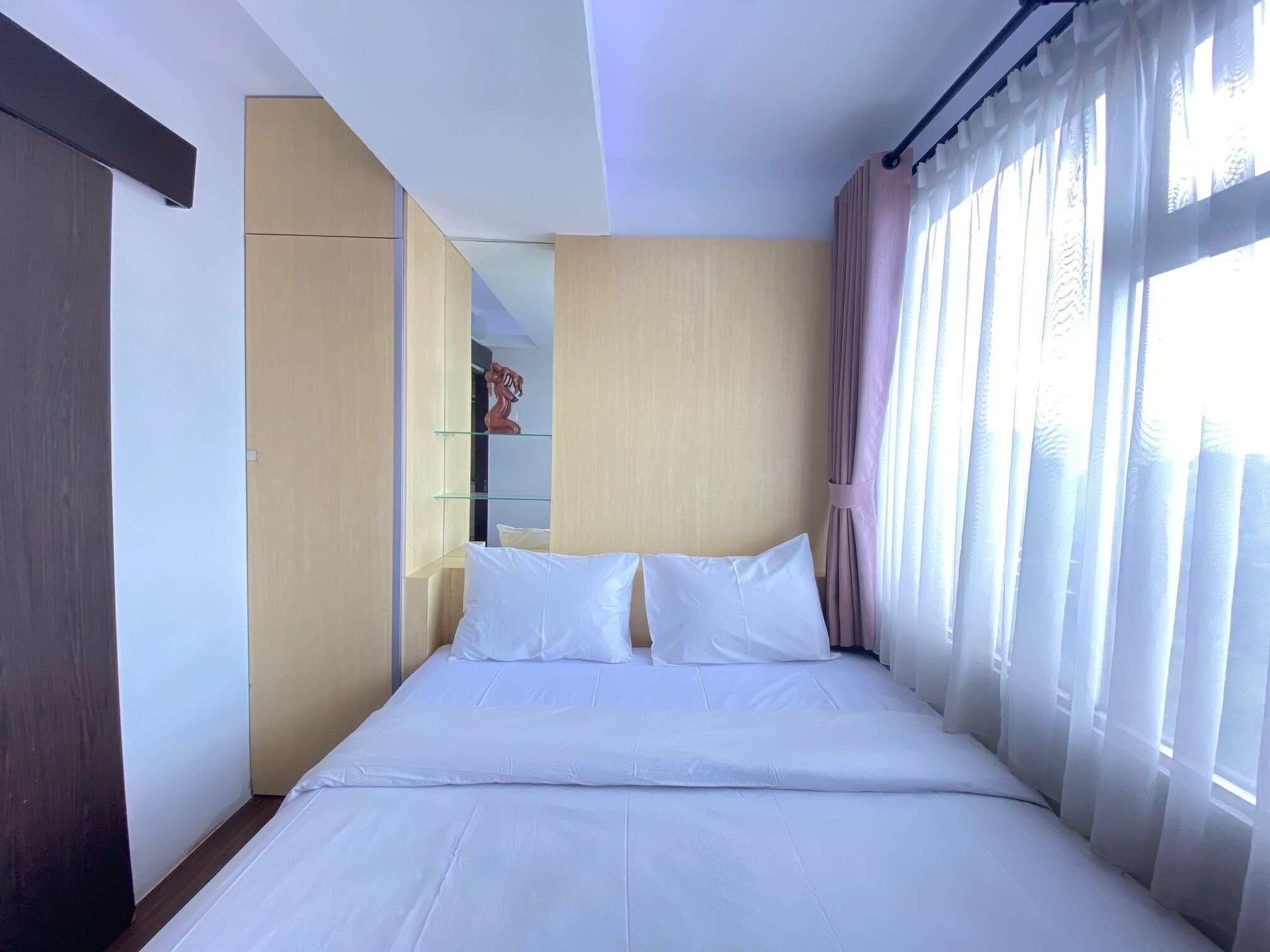 Une chambre d’hôtel avec un lit, une armoire, une étagère ornée d’un objet décoratif et des rideaux légers à la fenêtre.