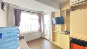 A hotel room with a large water dispenser, a flat-screen TV, wooden shelving, and a sliding glass door leading to a balcony. - Quite 2Br Apartment Ac In Living Room At The Jarrdin Cihampelas (Bandung)