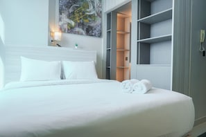 A neatly made bed with white linens and two rolled towels, a bedside lamp, and a painting on the wall. - Great Choice Deluxe Studio Apartment At Capitol Park Residence (Jakarta)