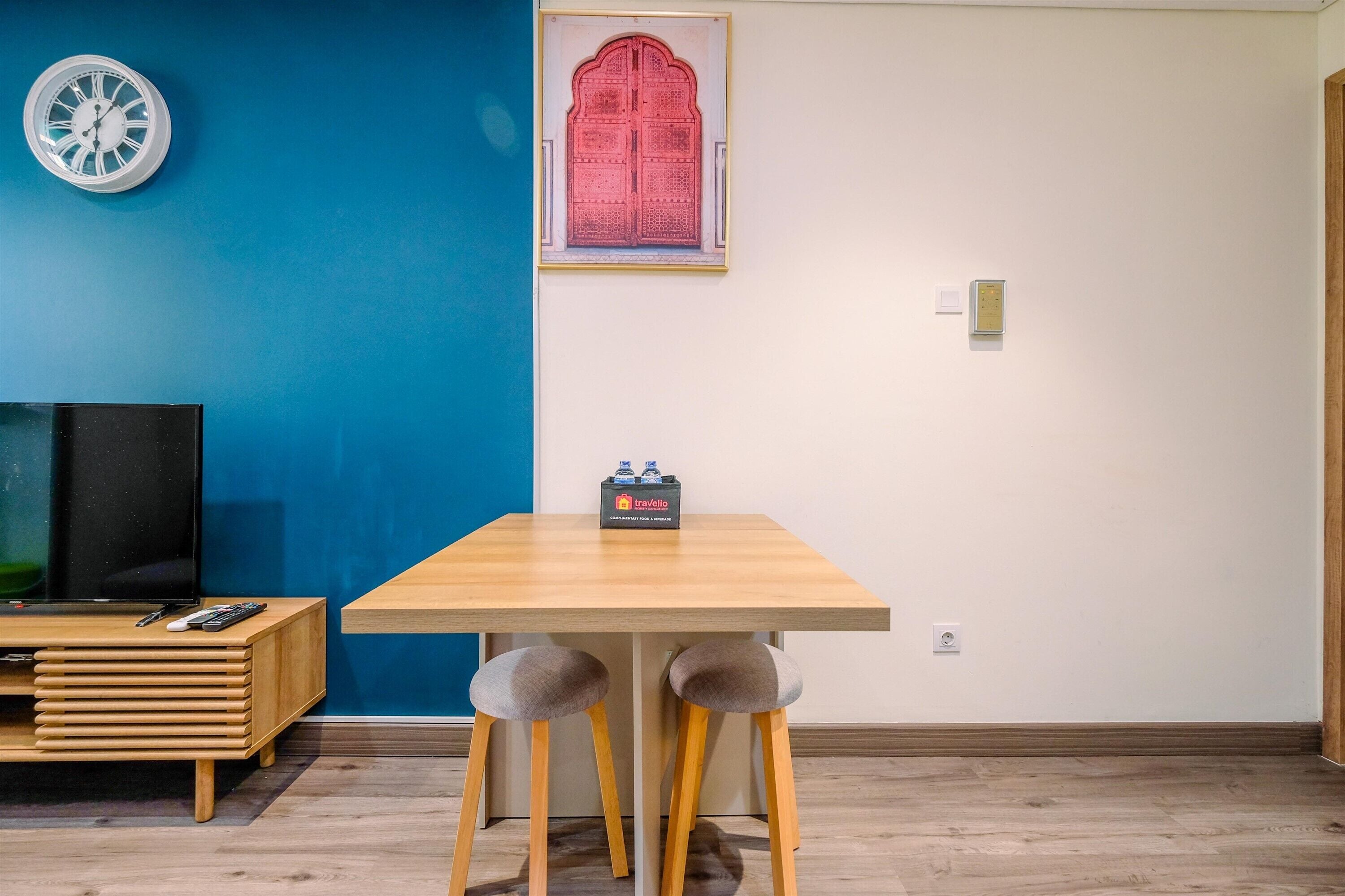A room with a blue wall, a wooden table, two grey stools, a framed red door, a clock, and a TV on a wooden stand.