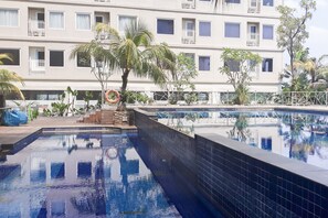 A swimming pool with tiled walls, surrounded by a fence and palm trees, with a multi-story building in the background.