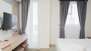 A hotel room with a wooden desk, two stools, a flat-screen TV, a bed with a rolled white towel, and grey curtains. - Brand New And Good Studio At Bintaro Icon Apartment (South Tangerang)