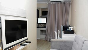 Um espaço compacto para viver, com televisão de tela plana, sofá cinza, cozinha com micro-ondas e cortina.