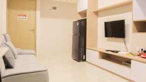 A modern living room with a sofa, a flat-screen TV mounted on the wall, a built-in wooden cabinet, and a refrigerator. - Great Choice 2Br Apartment At Meikarta (Cikarang)