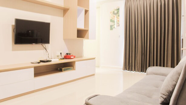 A modern living room with a flat-screen TV mounted on the wall, a wooden entertainment unit, a grey sofa, and a sliding glass door leading to a balcony. - Great Choice 2Br Apartment At Meikarta (Cikarang)